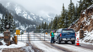 Three hikers with backpacks study a map beside a parked SUV at a snow-dusted mountain road gate, blocked for seasonal closure, with pine trees and misty foothills in the background.